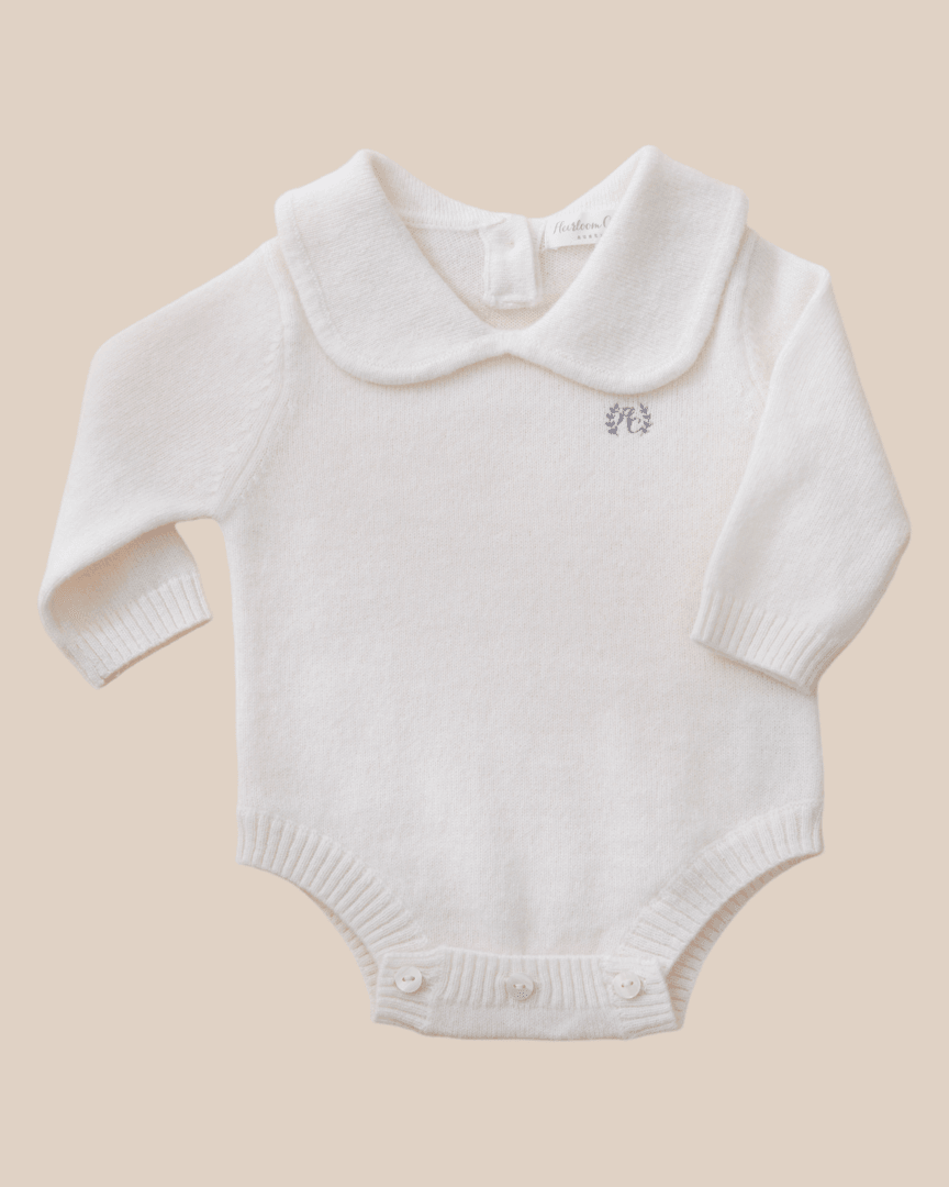 A white cashmere baby onesie with a Peter Pan collar, ribbed cuffs, and mother-of-pearl buttons.