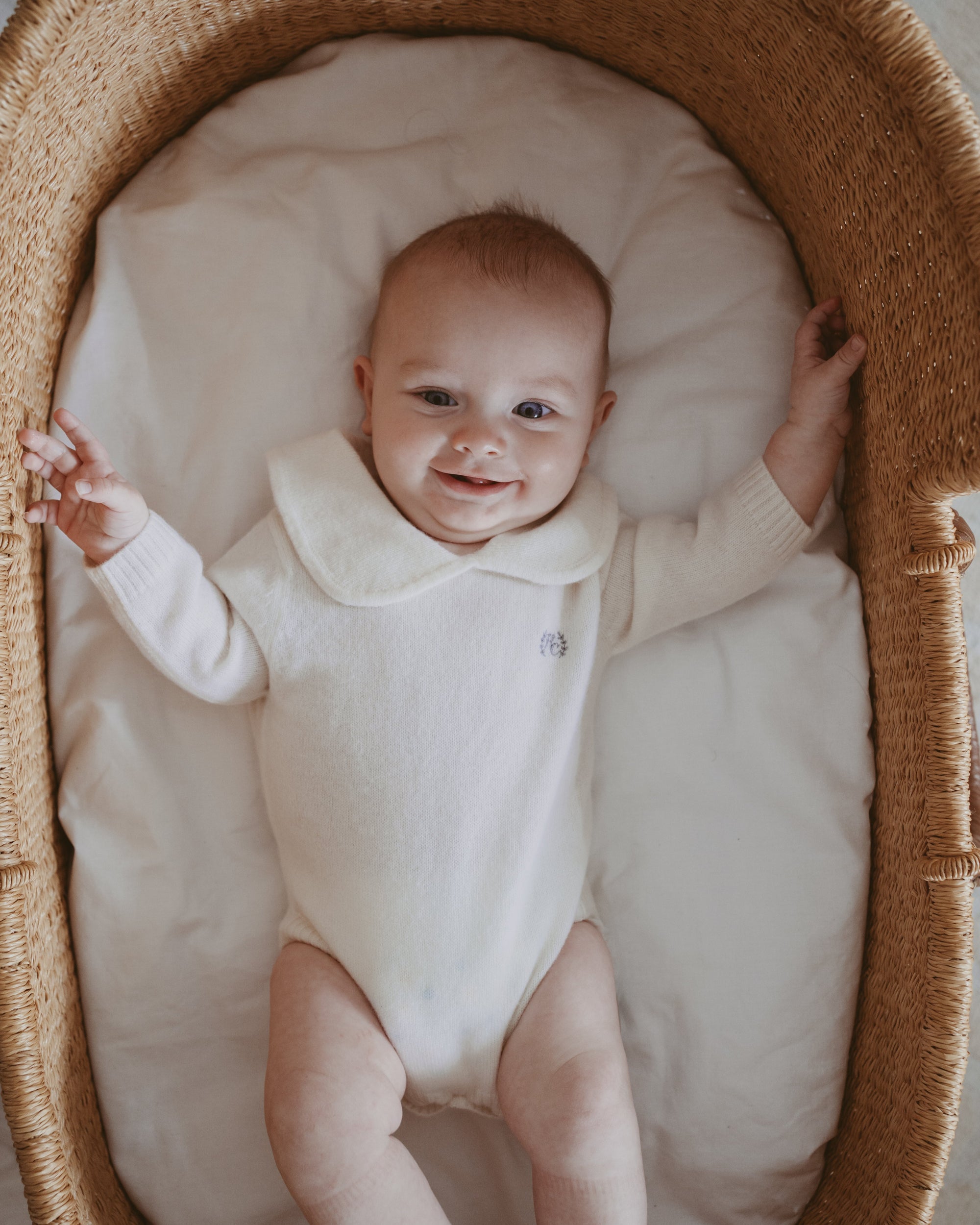 Baby in a cashmere onesie lying in a wicker crib with a soft white mattress.